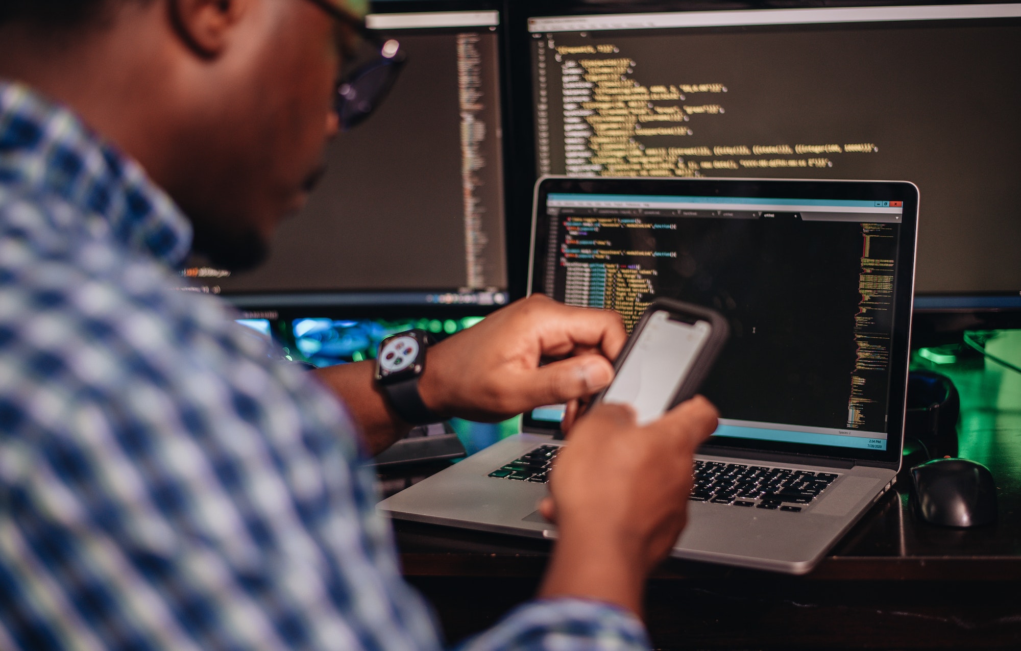 African American man sitting at computer developer software while using smartphone